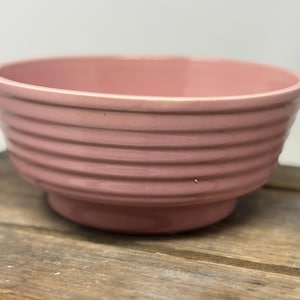Könnte beinhalten: Eine rosa Keramikschale mit gerippter Außenseite. Die Schale hat einen abgerundeten Boden und eine weite Öffnung. Die Farbe ist ein gedämpftes Rosa, und die Schale scheint aus Keramik zu sein. Die Schale steht auf einer Holzoberfläche.