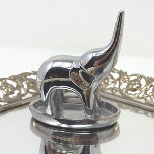 May include: A polished chrome elephant figurine standing on a small oval base. The elephant has a smooth, reflective surface and its trunk is raised upward. The figurine is positioned on a reflective surface with a decorative metal border.