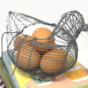 Op de afbeelding: Een draadmand in de vorm van een kip, gemaakt van dun grijs metaaldraadgaas. De mand bevat vier bruine kippeneieren, die zichtbaar zijn door de open draadstructuur van de kipvormige houder.