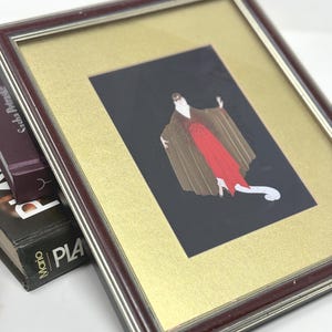 May include: Framed art print depicting a figure in a red dress and brown cape against a black backdrop. The artwork is set within a gold mount and a dark brown wooden frame with a silver inner edge. Books are visible in the background.