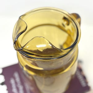 May include: A vintage amber glass pitcher with a curved spout and a handle. The pitcher has a rounded body and a smooth, glossy finish. The pitcher is sitting on a purple surface.