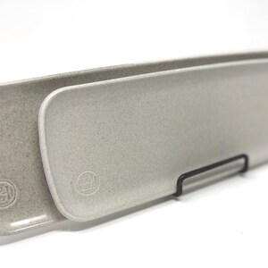 May include: Two elongated, rectangular ceramic serving trays in a speckled light gray finish. Each tray features rounded corners and a small, circular embossed logo stamp near one end. The trays are positioned upright on a thin black metal stand.