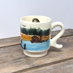 May include: A cream-colored ceramic mug featuring a hand-painted scene of a brown boat on blue water, a tan shoreline, and dark green trees under a light sky. The mug has a distinctively curved handle and a circular stamp inside the rim.