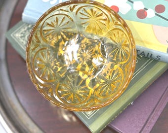 Könnte beinhalten: Eine runde, bernsteinfarbene Glasschale mit einem gepressten Muster aus strahlenförmigen Sternen- oder Sonnenmotiven auf der Oberfläche. Die Schale hat einen gewellten Rand und eine durchscheinende Beschaffenheit mit geometrischem Design.