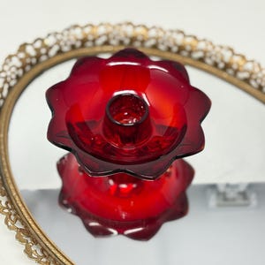 May include: A vibrant red glass candle holder with a flower-like design. The candle holder sits on a mirrored surface, reflecting its shape. The mirror is framed by a gold-colored, ornate, oval-shaped frame. The candle holder is a decorative item.