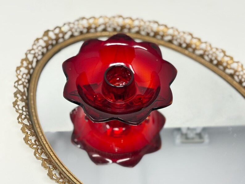 May include: A vibrant red glass candle holder with a flower-like design. The candle holder sits on a mirrored surface, reflecting its shape. The mirror is framed by a gold-colored, ornate, oval-shaped frame. The candle holder is a decorative item.