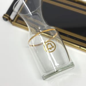 May include: A clear glass carafe with a gold band and a gold bell emblem. The carafe has a curved shape and is set against a black and gold decorative tray.