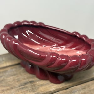 May include: A maroon-colored, oval-shaped ceramic planter with a glossy finish. The planter features a decorative, rope-like border and a pedestal base. The interior is a lighter shade of pink. The planter is sitting on a wooden surface.
