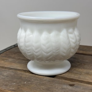 May include: A white, vintage-style planter with a rounded body and a pedestal base. The exterior features a textured, leaf-like pattern. The opening is wide, and the planter is made of opaque glass. This decorative piece is suitable for small plants or floral arrangements.