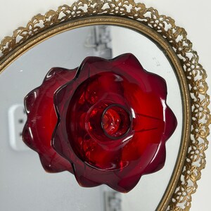 May include: A ruby red glass candle holder with a flower-like design, reflected in an oval mirror with a gold filigree frame. The candle holder has a central well for the candle and a scalloped edge.
