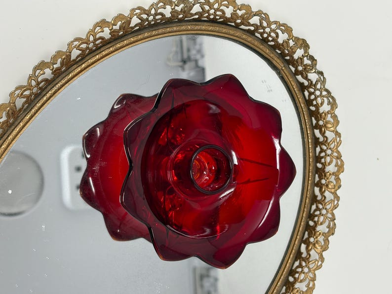May include: A ruby red glass candle holder with a flower-like design, reflected in an oval mirror with a gold filigree frame. The candle holder has a central well for the candle and a scalloped edge.