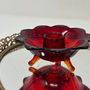 May include: A vintage red glass candle holder with a flower-shaped design. The candle holder has three small legs and a central cup to hold a candle. The glass is a deep red color, and the piece is reflected in the mirror below.