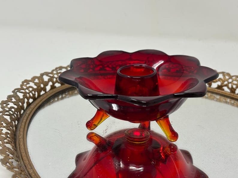 May include: A vintage red glass candle holder with a flower-shaped design. The candle holder has three small legs and a central cup to hold a candle. The glass is a deep red color, and the piece is reflected in the mirror below.