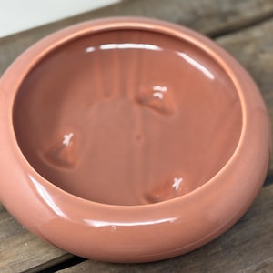 Puede incluir: Un cuenco de cerámica redondo de color terracota con un acabado liso y brillante. El interior presenta tres hendiduras triangulares distintas y elevadas en la base, creando un sutil patrón texturizado dentro del recipiente circular.