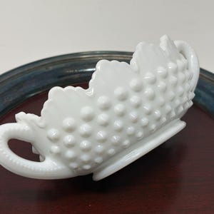 Puede incluir: Un plato de vidrio blanco con diseño de hobnail, con borde festoneado y dos asas curvas. El plato está decorado con pequeñas protuberancias en relieve y se encuentra sobre una superficie de madera oscura.