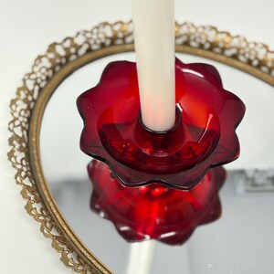 May include: A red glass candle holder with a flower-like design, holding a white candle. The candle holder sits on a gold-rimmed, mirrored tray. The candle holder is a deep, rich red color, and the tray has an ornate, vintage design.