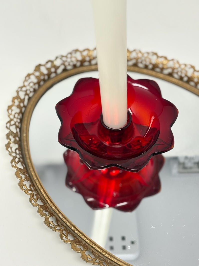 May include: A red glass candle holder with a flower-like design, holding a white candle. The candle holder sits on a gold-rimmed, mirrored tray. The candle holder is a deep, rich red color, and the tray has an ornate, vintage design.
