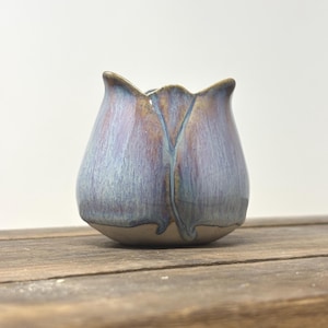 Könnte beinhalten: Ein kleines Keramikgefäß in Tulpenform mit gewelltem Rand. Die Außenseite ist mit einer reaktiven Glasur in gemischten Tönen von Lavendel, sanftem Blau und hellbraunen Akzenten versehen, mit einem abgerundeten Boden und einer vertikalen Naht.