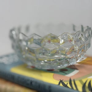 Peut inclure: Un bol en verre transparent facetté avec un motif géométrique. Le bol repose sur un livre avec une couverture bleue et des illustrations colorées. Les bords du bol ont un design festonné.