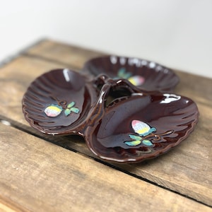 May include: A dark brown ceramic serving dish with three shell-shaped compartments and a central handle. Each section features a hand-painted motif of a small fruit with green leaves, rendered in yellow, pink, and white tones on the glossy surface.