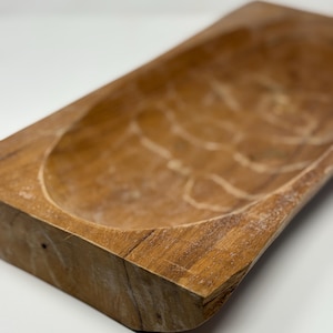 May include: A rectangular, wooden dough bowl with a natural finish. The bowl has a carved interior and a rustic, weathered appearance. The wood grain is visible, adding to its charm. Perfect for home decor or serving.
