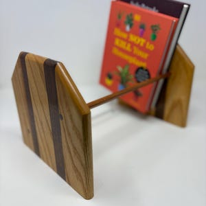 Puede incluir: Soporte de libro de madera con un diseño a rayas marrón claro y marrón oscuro. El soporte contiene dos libros, uno con una cubierta roja y el texto "How NOT to Kill Your Houseplant". El soporte es de madera y tiene un diseño sencillo y moderno.