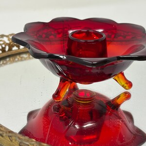 May include: A vintage red glass candle holder with a flower-shaped design. The candle holder has three small feet and a central cup to hold a candle. The glass is a deep red color, and the piece is likely from the mid-century.