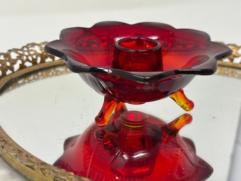 May include: A vintage red glass candle holder with a flower-shaped design. The candle holder has three small feet and a central cup to hold a candle. The glass is a deep red color, and the piece is likely from the mid-century.