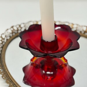 May include: A ruby red glass candle holder with a flower-shaped design. The candle holder is on a mirrored surface, reflecting its shape. A white candle is inserted in the center. The candle holder sits on a gold-colored, ornate tray.