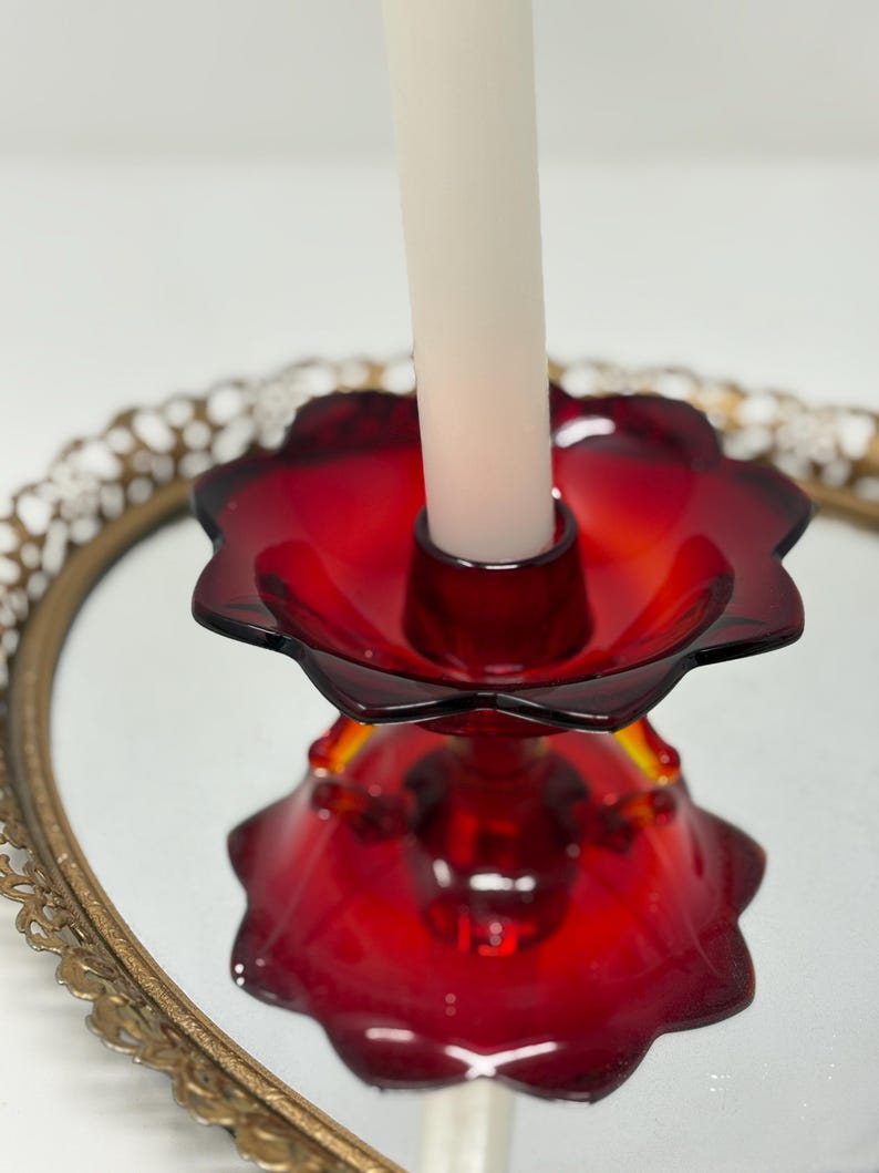May include: A ruby red glass candle holder with a flower-shaped design. The candle holder is on a mirrored surface, reflecting its shape. A white candle is inserted in the center. The candle holder sits on a gold-colored, ornate tray.