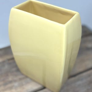 May include: A pale yellow ceramic vase with a rectangular, slightly tapered silhouette and a smooth, glossy finish. The vase features a wide, open top with clean, straight edges and a solid, uniform color throughout its entire exterior surface.