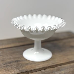 May include: A white glass dessert bowl with a ruffled edge. The bowl sits on a pedestal base. The rim has a scalloped design. The bowl is on a wooden surface.