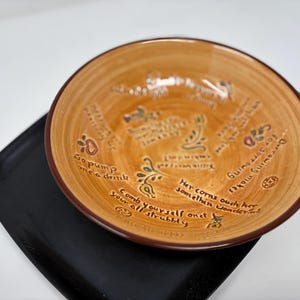 May include: A round, tan ceramic bowl with a brown rim, resting on a black square tray. The bowl features handwritten text and decorative floral elements in various colors. The text includes phrases like "Go pump me a drink" and "Comb yourself".
