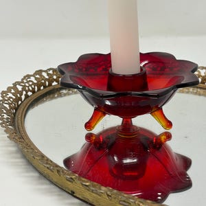 Può includere: Un portacandele vintage in vetro rosso con una parte superiore a forma di fiore e tre piedini giallo-arancio. Una candela bianca è inserita al centro. Il portacandele è su un vassoio a specchio con bordo in filigrana dorata.