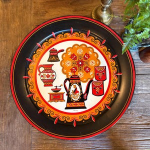 May include: A vintage black metal serving tray with a red and orange floral design. The tray features a central white circle with coffee-themed illustrations, including a coffee pot, sugar and coffee cans, and a coffee grinder. The tray's border is decorated with orange and red swirls.