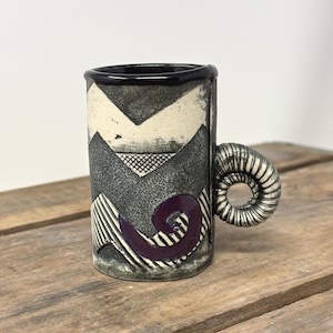 May include: A ceramic mug with a black rim and a textured, cylindrical body featuring a gray and white chevron pattern, a purple spiral accent, and a distinct, coiled handle that resembles a ram's horn, finished with a matte, speckled surface.