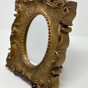 May include: A gold-colored, ornate, oval-shaped mirror with a decorative frame. The frame has a detailed, intricate design with a floral pattern.