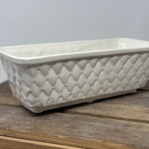 May include: An off-white ceramic rectangular planter with a textured, woven design. The planter has a slightly flared rim and is placed on a wooden surface. Suitable for indoor or outdoor use, it adds a touch of elegance to any space.