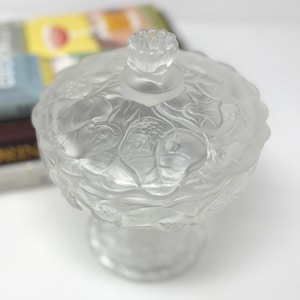 Puede incluir: Un cuenco para dulces de vidrio esmerilado transparente con tapa, que presenta un patrón en relieve de hojas y flores de nenúfar. La tapa está rematada con un pequeño mango esculpido en forma de flor y descansa sobre una base.