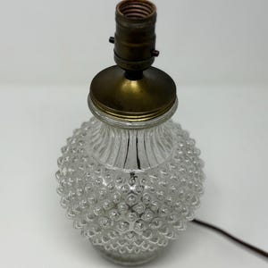 Puede incluir: Una lámpara de mesa de vidrio transparente con un patrón texturizado en forma de burbujas en la base. La lámpara tiene una parte superior de metal color latón y un cable eléctrico marrón. La lámpara está diseñada para una bombilla estándar.