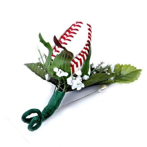 May include: A baseball-themed boutonniere with a white baseball with red stitching, white flowers, and green leaves. The boutonniere is attached to a green stem with a silver pin.