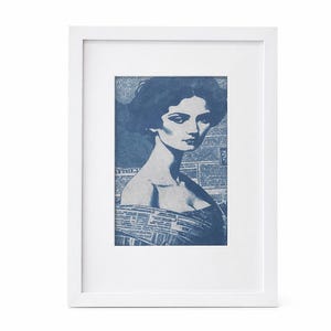 Puede incluir: Una impresión enmarcada con un retrato en tonos azules de una mujer. La obra de arte se encuentra sobre un fondo de impresión de periódico, con la imagen de la mujer en primer plano. El marco blanco crea un contraste limpio.