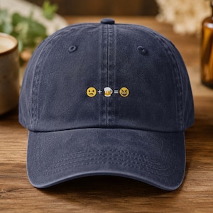 May include: A navy blue baseball cap featuring an embroidered graphic on the front center. The design consists of a sad face emoji, a plus sign, a beer mug icon, an equals sign, and a laughing emoji, all stitched in white, yellow, and brown.