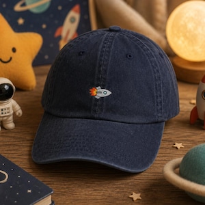 May include: A navy blue baseball cap featuring a small, embroidered graphic of a white rocket ship with orange and yellow flames trailing behind it. The cap has a structured crown, a curved brim, and a classic six-panel design with top stitching.
