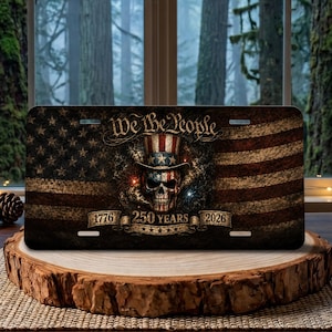 Puede incluir: Placa de matrícula con un diseño de bandera estadounidense y una calavera con un sombrero patriótico. El texto "We the People" está en la parte superior, con "1776 250 YEARS 2026" en la parte inferior. La placa se muestra sobre una superficie de madera.