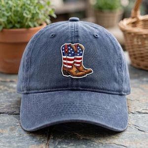 Puede incluir: Gorra de béisbol azul marino con un parche que representa botas de vaquero con los colores de la bandera estadounidense. Las botas son marrones con rayas y estrellas rojas, blancas y azules. La gorra es de material suave y desgastado.