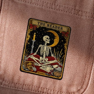 Puede incluir: Una carta de tarot rectangular con un esqueleto en posición meditativa, con una luna creciente y una vela. La carta está etiquetada como "THE READER" en la parte superior y "BOOKS" en la inferior, sobre un fondo oscuro con detalles dorados.