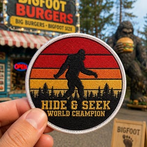 May include: A circular embroidered patch featuring a black silhouette of Bigfoot walking against a sunset background of red, orange, and yellow horizontal stripes above a black forest. The text reads HIDE & SEEK WORLD CHAMPION in gold.