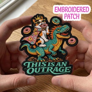 May include: Embroidered patch featuring a cat wearing a crown and sunglasses, riding a dinosaur and eating a donut. The patch has the text "THIS IS AN OUTRAGE" in green. The patch is black with colorful accents of orange, blue, and pink.