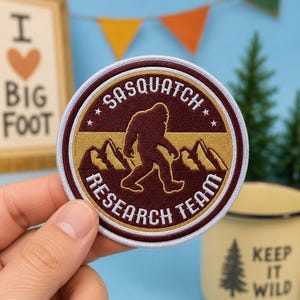 May include: A circular embroidered patch featuring a maroon and gold design. The center depicts a Sasquatch silhouette against mountain peaks. The text reads SASQUATCH RESEARCH TEAM in white lettering, accented by four small white stars.